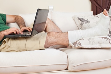 A middle aged man sitting on the couch with a broken leg and a laptop working from home