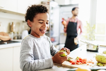 A young boy laughing while he helps his dad prepare lunch by peeling some fruit and veg.